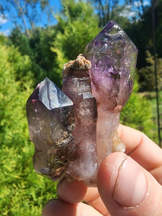 May include: Amethyst cluster featuring three main points, exhibiting a captivating blend of deep purple and smoky gray hues. The crystals showcase varying degrees of translucence, with some areas appearing more transparent than others.  The cluster is naturally formed with visible matrix material at the base.  The amethyst points are sharply defined, displaying natural crystal formations.  This raw amethyst specimen is a unique addition to any mineral collection.